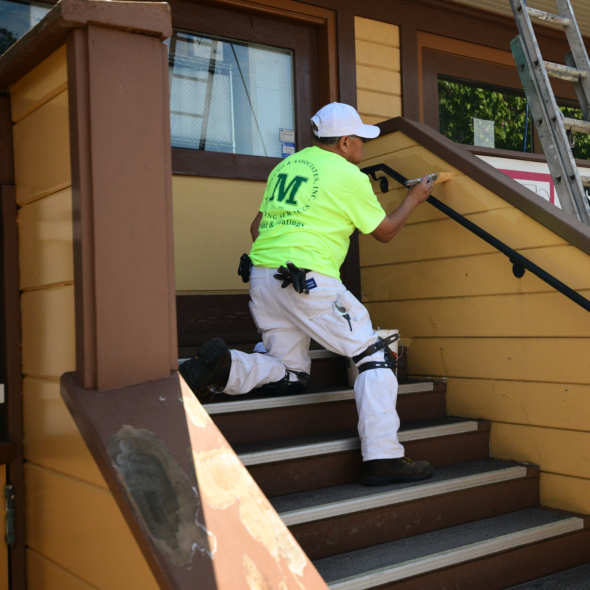 Man in neon yellow shirt and white pants wearing a white hat painting the exterior of a building standing on a flight of stairs.