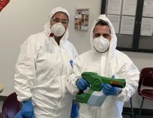 two people one male and one female dressed with a hazmat suit. The male is holding an electrostatic disinfection sprayer.
