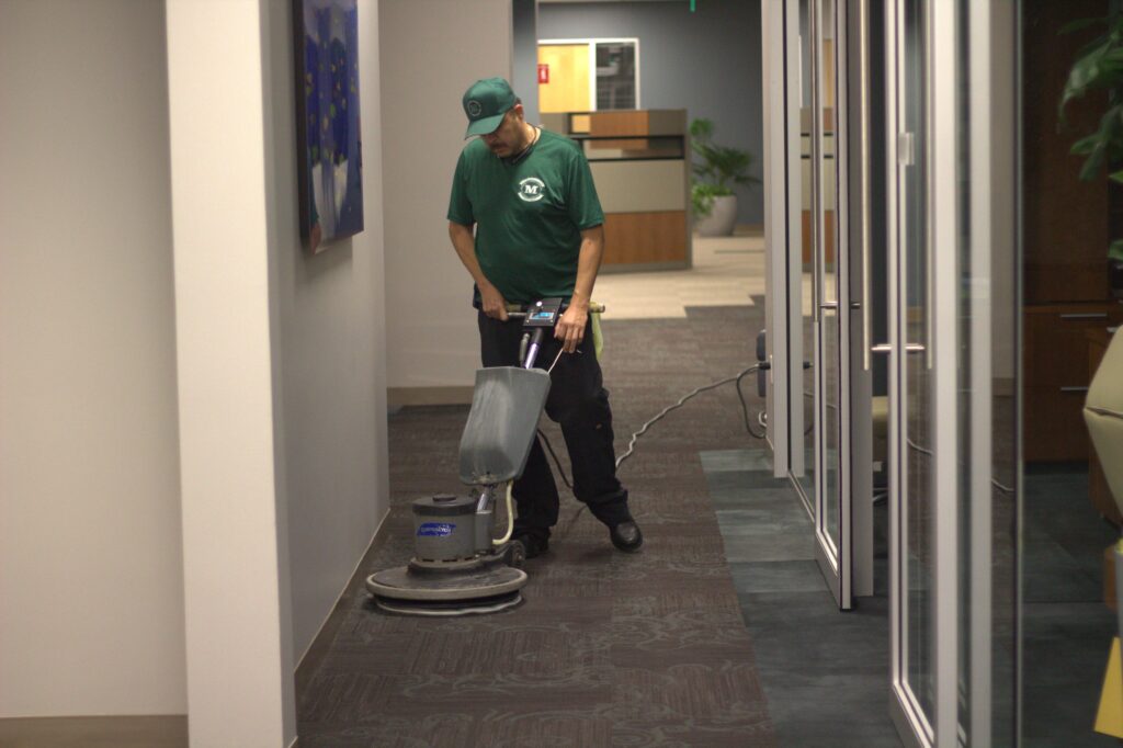 Man in green shirt, black pants, and green hat. Using a rotary floor scrubbing machine on carpet.