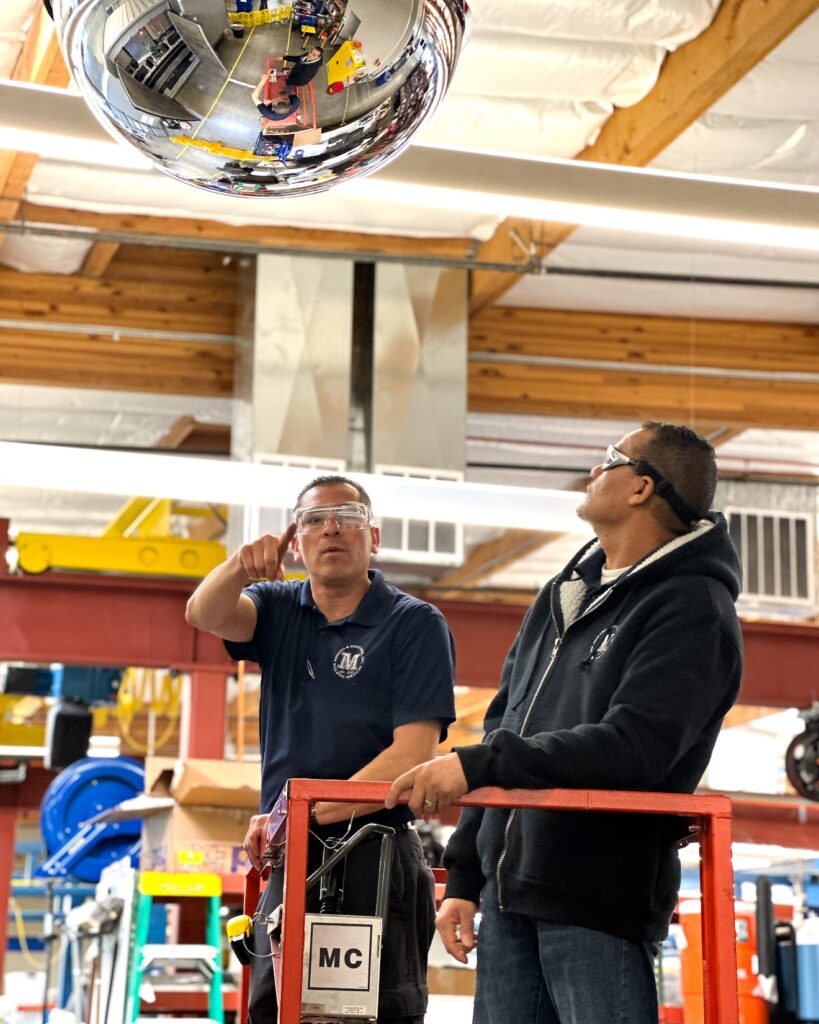 two male Moreno employee's working in a warehouse standing on top of a lift. One employee is pointing as the other inspects his work.