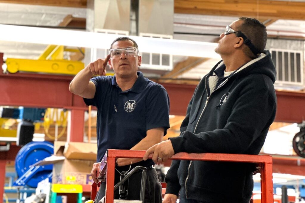 two male Moreno employee's working in a warehouse standing on top of a lift. One employee is pointing as the other inspects his work.